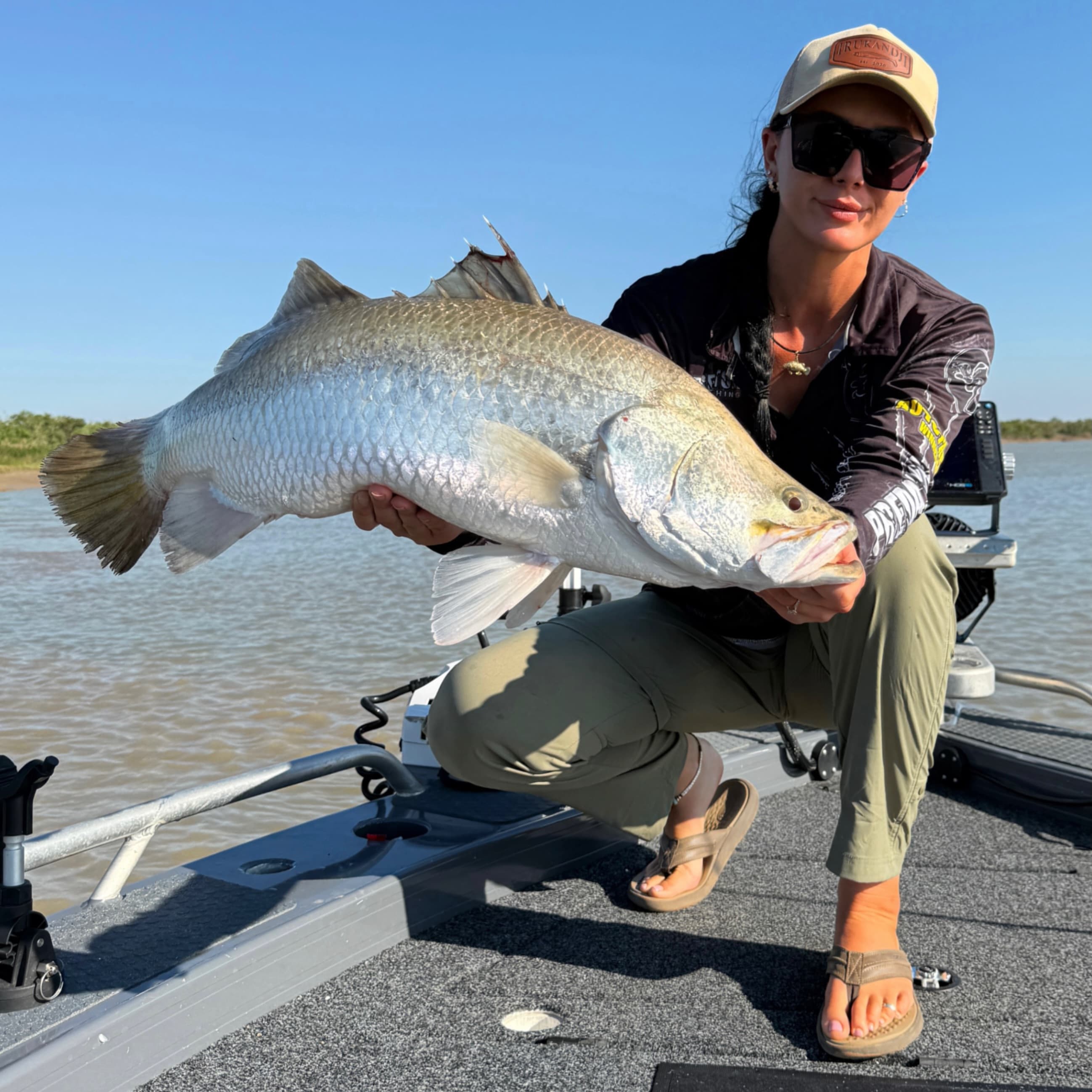 Sarah Vandy crouched on the boat holding a large barramundi at sunrise
